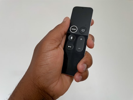 A hand holds a sleek, black remote control featuring several buttons including menu, play/pause, and volume controls. The remote has a modern design with a glossy finish.