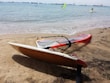 A windsurfing board with an orange sail rests on a sandy beach. The sea is calm with a few visible waves. In the background, several people are windsurfing on the water, and numerous ships can be seen on the horizon.