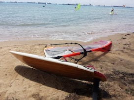 A windsurfing board with an orange sail rests on a sandy beach. The sea is calm with a few visible waves. In the background, several people are windsurfing on the water, and numerous ships can be seen on the horizon.
