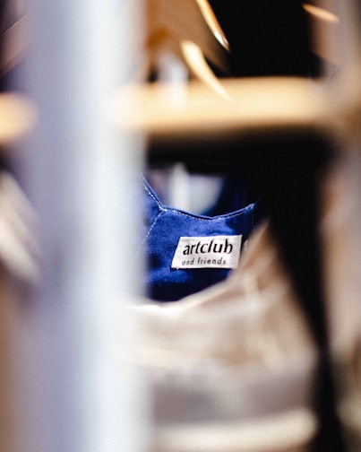 A clothing label with the text 'artclub and friends' is sewn onto a blue fabric. The image is focused on the label with blurred foreground and background, suggesting the photograph was taken in a clothing store or closet environment.