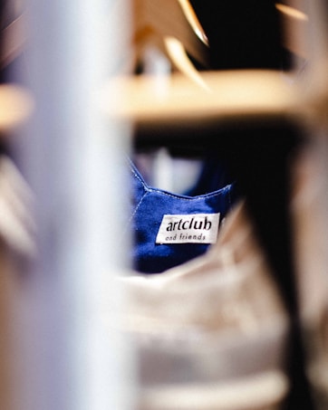 A clothing label with the text 'artclub and friends' is sewn onto a blue fabric. The image is focused on the label with blurred foreground and background, suggesting the photograph was taken in a clothing store or closet environment.