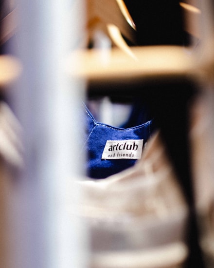 A clothing label with the text 'artclub and friends' is sewn onto a blue fabric. The image is focused on the label with blurred foreground and background, suggesting the photograph was taken in a clothing store or closet environment.