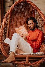 A man with glasses sits comfortably in a woven hanging chair while reading an orange book titled 'Think and Grow'. He wears a bright red shirt and white pants, and the setting exudes a relaxed, cozy atmosphere.