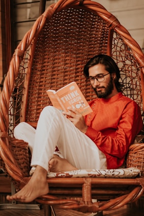 A man with glasses sits comfortably in a woven hanging chair while reading an orange book titled 'Think and Grow'. He wears a bright red shirt and white pants, and the setting exudes a relaxed, cozy atmosphere.