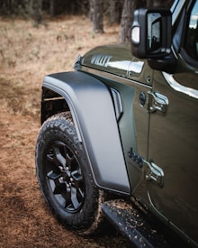 A close-up of the front side of a rugged off-road vehicle, focusing on the wheel, fender, and side mirror. The car body is olive green with visible dirt marks, reflecting its outdoor use. The surroundings include a grassy and wooded area, suggesting an off-road setting.