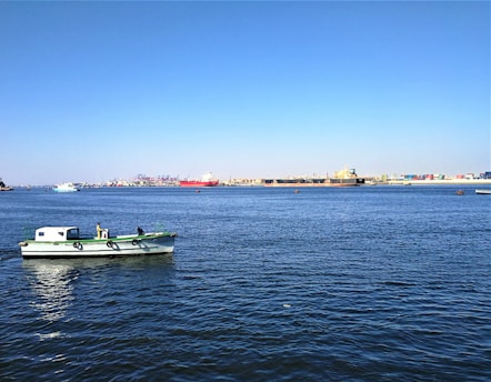 A small green and white boat with two people on it is on calm blue water. In the background, there are large container ships and port facilities with various cranes and stacked containers.