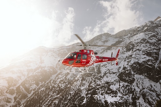 A red rescue helicopter hovers in the foreground against a backdrop of snowy mountains. The aircraft features distinct white markings and a medical cross, indicating its function as an emergency or rescue vehicle. The mountain range is partially illuminated by sunlight, creating dramatic contrasts of shadow and light across the terrain.