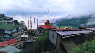 A scenic view of a hillside village featuring colorful buildings with metal and red roofs. A central structure with a red roof and prayer flags is prominently positioned, surrounded by residential buildings. Green hills rise in the background, partially obscured by misty clouds, creating a serene atmosphere.
