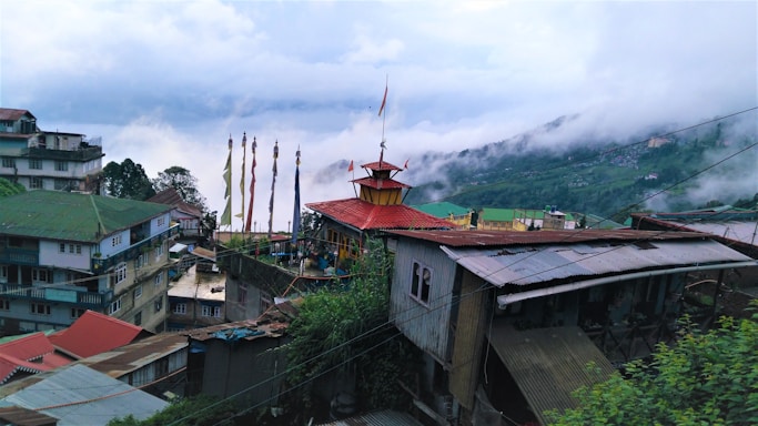 A scenic view of a hillside village featuring colorful buildings with metal and red roofs. A central structure with a red roof and prayer flags is prominently positioned, surrounded by residential buildings. Green hills rise in the background, partially obscured by misty clouds, creating a serene atmosphere.