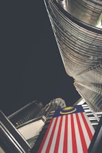 A dramatic upward perspective showcases a towering skyscraper with reflective glass and steel architecture. A prominent Malaysian flag is draped vertically, adding color contrast to the modern structure. The night sky envelops the scene, suggesting a city environment.