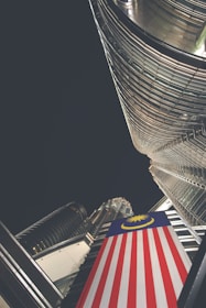 A dramatic upward perspective showcases a towering skyscraper with reflective glass and steel architecture. A prominent Malaysian flag is draped vertically, adding color contrast to the modern structure. The night sky envelops the scene, suggesting a city environment.