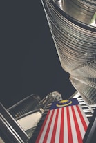 A dramatic upward perspective showcases a towering skyscraper with reflective glass and steel architecture. A prominent Malaysian flag is draped vertically, adding color contrast to the modern structure. The night sky envelops the scene, suggesting a city environment.