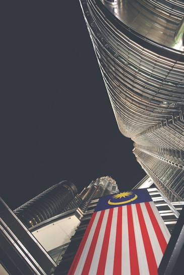 A dramatic upward perspective showcases a towering skyscraper with reflective glass and steel architecture. A prominent Malaysian flag is draped vertically, adding color contrast to the modern structure. The night sky envelops the scene, suggesting a city environment.