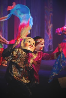 Two performers wear traditional and colorful costumes, including vivid masks with painted expressions. Flowing fabric in bright hues of blue, pink, and orange adds movement. The scene takes place on a dimly lit stage with intricate patterns in the background.