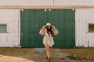 woman in white and brown floral dress wearing white hat standing in front of green wooden