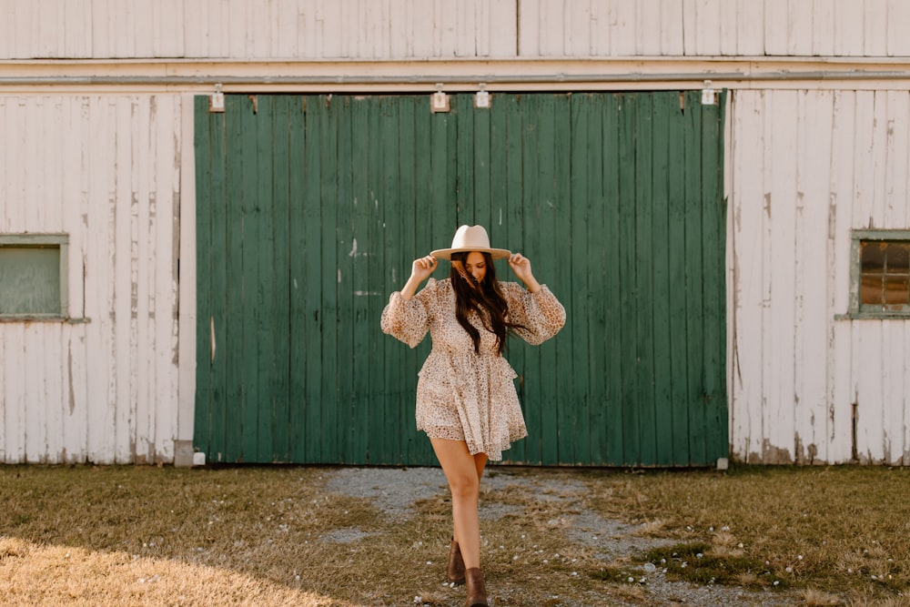 woman in white and brown floral dress wearing white hat standing in front of green wooden