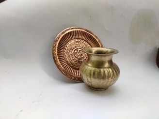 A brass or bronze pot with a rounded body is placed next to an intricately designed, circular metallic plate with a central emblem. The background is plain and light.