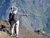 Two individuals stand on a rocky terrain with one person pointing a trekking pole toward the mountainous landscape in the background. They are dressed in outdoor gear, including beanies and jackets, and one carries a backpack with polka dots.
