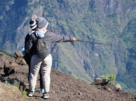 Two individuals stand on a rocky terrain with one person pointing a trekking pole toward the mountainous landscape in the background. They are dressed in outdoor gear, including beanies and jackets, and one carries a backpack with polka dots.