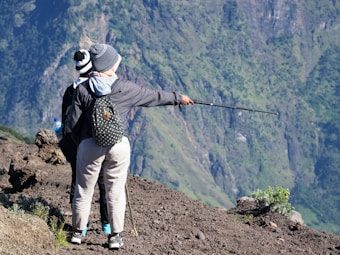 Two individuals stand on a rocky terrain with one person pointing a trekking pole toward the mountainous landscape in the background. They are dressed in outdoor gear, including beanies and jackets, and one carries a backpack with polka dots.