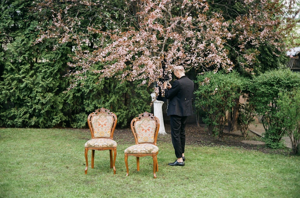 man in black suit standing near brown wooden armchair