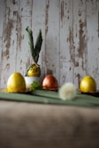 A rustic background features decorative eggs, two of which are yellow, and one is a reddish color. One of the yellow eggs is dressed with green fabric tied into what resembles ears, creating a character-like appearance. The eggs are placed on a green surface, with a blurred dandelion in the foreground and some greenery beside the eggs.