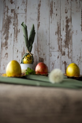 A rustic background features decorative eggs, two of which are yellow, and one is a reddish color. One of the yellow eggs is dressed with green fabric tied into what resembles ears, creating a character-like appearance. The eggs are placed on a green surface, with a blurred dandelion in the foreground and some greenery beside the eggs.