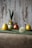 A rustic background features decorative eggs, two of which are yellow, and one is a reddish color. One of the yellow eggs is dressed with green fabric tied into what resembles ears, creating a character-like appearance. The eggs are placed on a green surface, with a blurred dandelion in the foreground and some greenery beside the eggs.