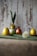 A rustic background features decorative eggs, two of which are yellow, and one is a reddish color. One of the yellow eggs is dressed with green fabric tied into what resembles ears, creating a character-like appearance. The eggs are placed on a green surface, with a blurred dandelion in the foreground and some greenery beside the eggs.