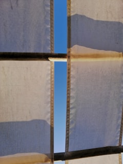 White fabric panels with visible seams are stretched across a structure, creating a partial canopy. Sunlight casts shadows and highlights on the fabric, revealing a clear blue sky through a slender gap between the panels.