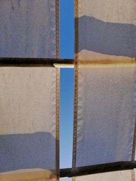 White fabric panels with visible seams are stretched across a structure, creating a partial canopy. Sunlight casts shadows and highlights on the fabric, revealing a clear blue sky through a slender gap between the panels.