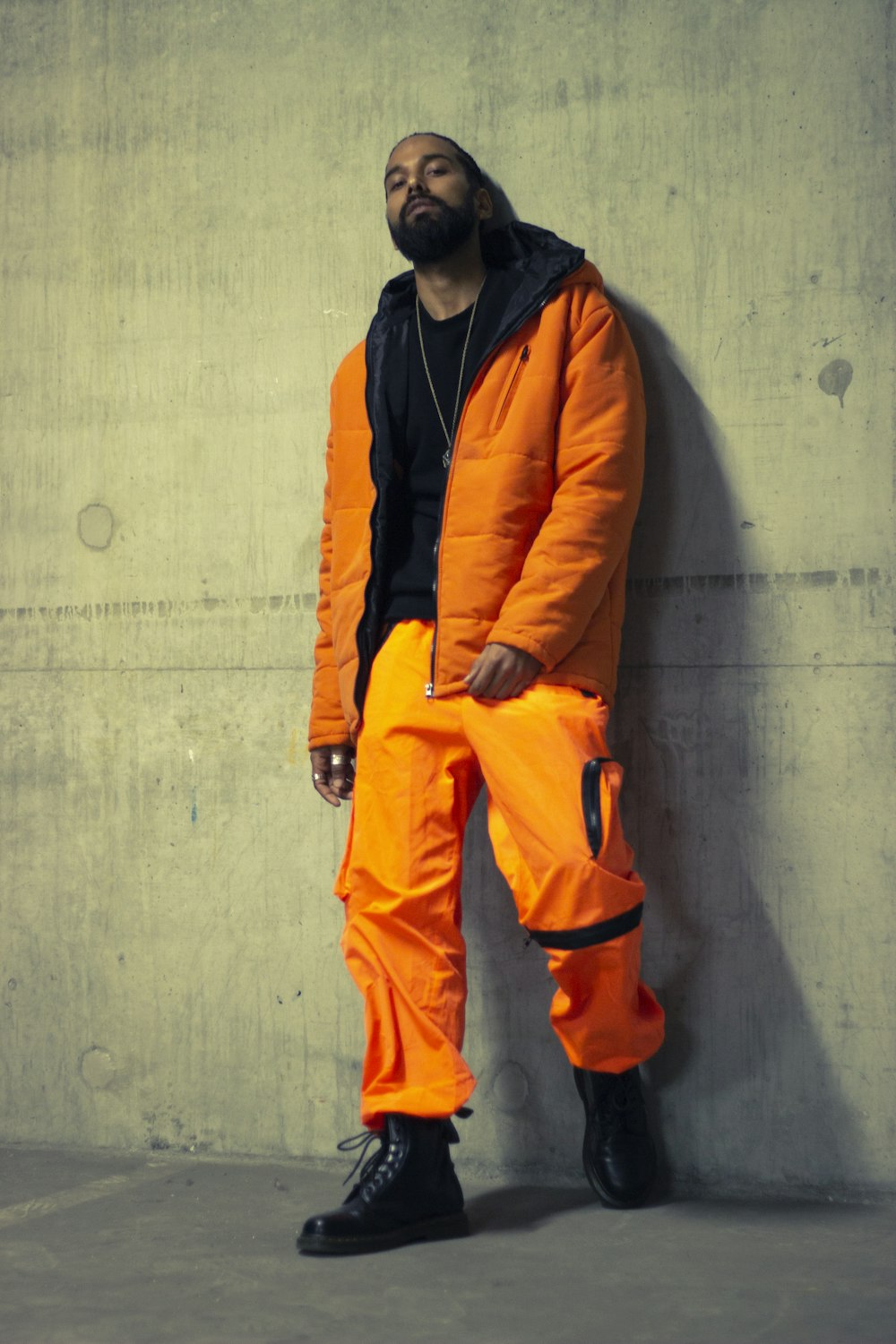 man in orange jacket leaning on gray wall