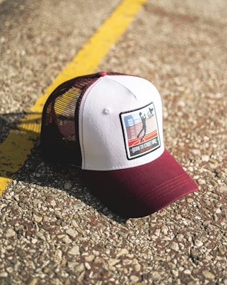 A baseball cap with a maroon brim and mesh back, featuring a patch with a basketball scene and the words 'Born to Street Ball'. The cap is resting on a textured concrete surface with a yellow line running through the background.