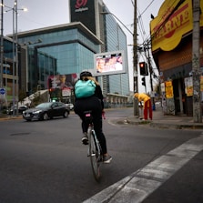 A cyclist wearing a light blue backpack rides along an urban street. The scene includes a large modern glass building with advertisements, a yellow corner store with a red sign, and a person dressed in red and yellow standing nearby. Traffic lights and cars are visible on the road.