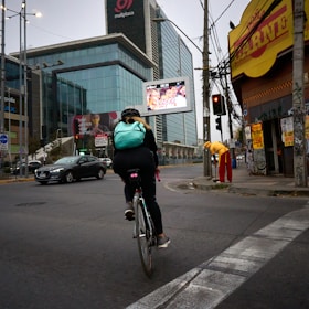 A cyclist wearing a light blue backpack rides along an urban street. The scene includes a large modern glass building with advertisements, a yellow corner store with a red sign, and a person dressed in red and yellow standing nearby. Traffic lights and cars are visible on the road.