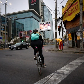 A cyclist wearing a light blue backpack rides along an urban street. The scene includes a large modern glass building with advertisements, a yellow corner store with a red sign, and a person dressed in red and yellow standing nearby. Traffic lights and cars are visible on the road.