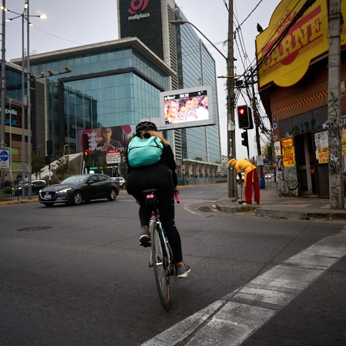 A cyclist wearing a light blue backpack rides along an urban street. The scene includes a large modern glass building with advertisements, a yellow corner store with a red sign, and a person dressed in red and yellow standing nearby. Traffic lights and cars are visible on the road.