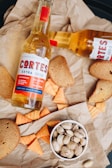 A composition featuring a bottle of beer labeled 'Cortes Extra Cerveza' alongside toasted bread slices, triangular corn snacks, and a bowl of pistachios, all arranged on crumpled brown paper.