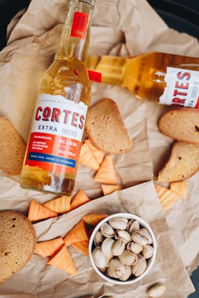 A composition featuring a bottle of beer labeled 'Cortes Extra Cerveza' alongside toasted bread slices, triangular corn snacks, and a bowl of pistachios, all arranged on crumpled brown paper.
