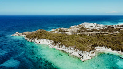 A scenic coastline with clear turquoise waters and a rugged, green hillside. Buildings are visible in the background, suggesting development on or near the coast. The horizon shows a vast expanse of deep blue ocean under a clear sky with a few clouds.
