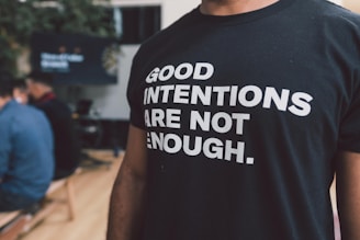 A person is wearing a black t-shirt with bold white text reading 'Good intentions are not enough.' The background features people sitting, possibly in a casual or meeting setting.