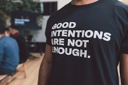 A person is wearing a black t-shirt with bold white text reading 'Good intentions are not enough.' The background features people sitting, possibly in a casual or meeting setting.