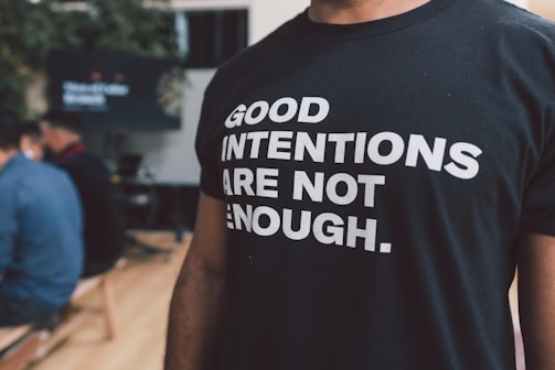 A person is wearing a black t-shirt with bold white text reading 'Good intentions are not enough.' The background features people sitting, possibly in a casual or meeting setting.