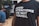 A person is wearing a black t-shirt with bold white text reading 'Good intentions are not enough.' The background features people sitting, possibly in a casual or meeting setting.