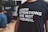A person is wearing a black t-shirt with bold white text reading 'Good intentions are not enough.' The background features people sitting, possibly in a casual or meeting setting.