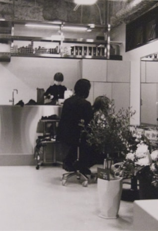 A hair salon with a person sitting on a chair, facing away, getting their hair styled or cut by a stylist. The salon has a modern and clean interior with a counter at the back, shelves with various products, and large plants in the foreground. There is another person in the background behind the counter, possibly an employee.