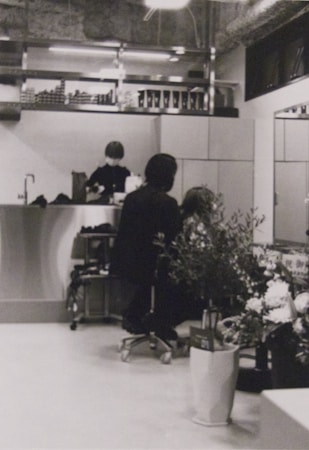 A hair salon with a person sitting on a chair, facing away, getting their hair styled or cut by a stylist. The salon has a modern and clean interior with a counter at the back, shelves with various products, and large plants in the foreground. There is another person in the background behind the counter, possibly an employee.