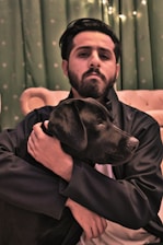 A person with a serious expression is sitting on a sofa, holding a black Labrador Retriever in a warm embrace. The background features green curtains with small patterns and some string lights, creating a cozy atmosphere.