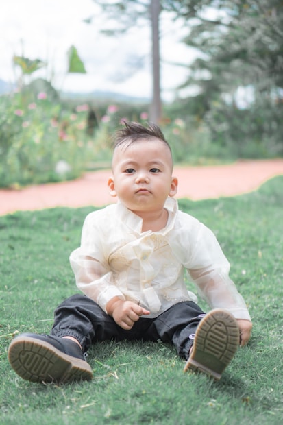 A young child sits on the grass in a garden setting, wearing a traditional white embroidered top and dark pants. The background features blurred greenery and flowers, highlighting a peaceful outdoor environment.