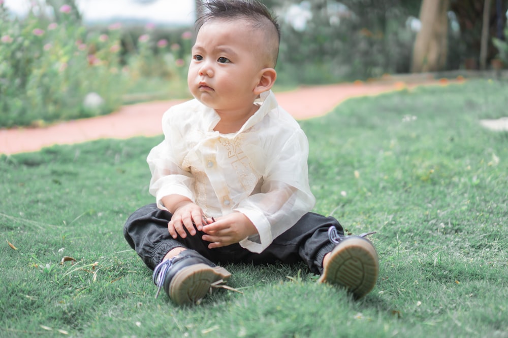 bebé con camisa de vestir blanca y pantalones negros sentado en la hierba verde durante el día